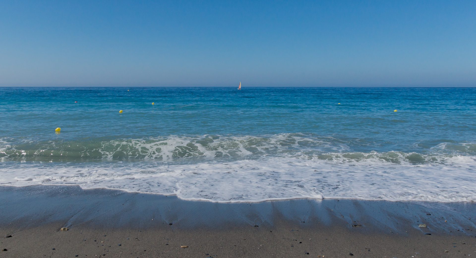 Mediterranean surf running up an Andalusian beach under a clear blue sky, a sail on the horizon